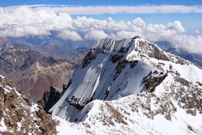 A wide-angle landscape of the jagged limestone peak of Carstensz Pyramid in Central Papua, showing the iconic vertical rock face and the rare tropical glaciers and snow patches near the summit under a clear blue sky
