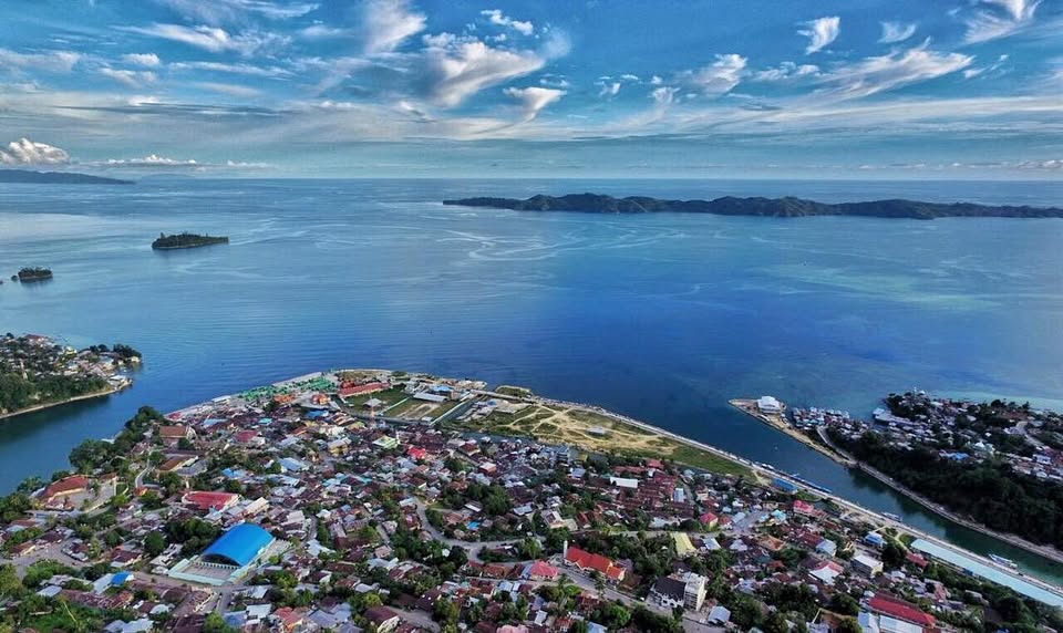"High-angle drone photograph of Fakfak City, showing colorful buildings nestled on a steep coastal hillside overlooking the blue waters of the Banda Sea, with numerous traditional longtail boats anchored near the shoreline."
