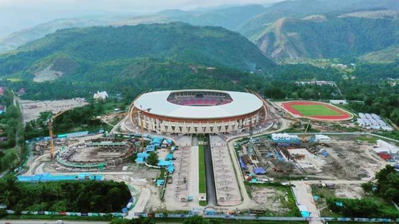 An aerial perspective of the Lukas Enembe Stadium in Papua, showcasing its modern, massive circular architecture with a distinctive roof design inspired by traditional Papuan motifs, surrounded by sports complex facilities and hills.