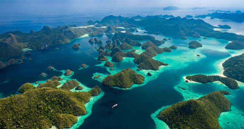 Aerial drone view of numerous lush green karst islands scattered across vibrant turquoise and deep blue ocean waters in Raja Ampat, Indonesia.