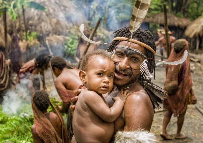 An indigenous Papuan woman smiling while carrying her baby in a traditional Noken bag woven from wood fiber, resting against her forehead.