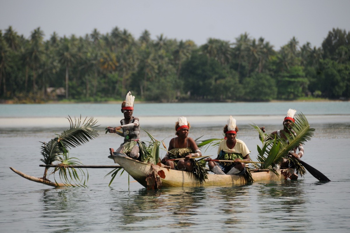 Four Melanesian men in traditional attire smiling and paddling a large outrigger canoe on clear turquoise ocean water near a tropical island coastline.