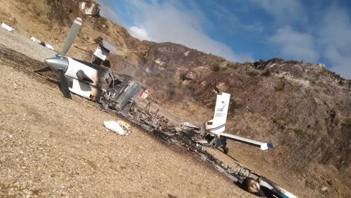 A charred and completely burned civilian perintis aircraft on a remote dirt runway, with only its tail section and propeller remaining intact amidst ash and debris.