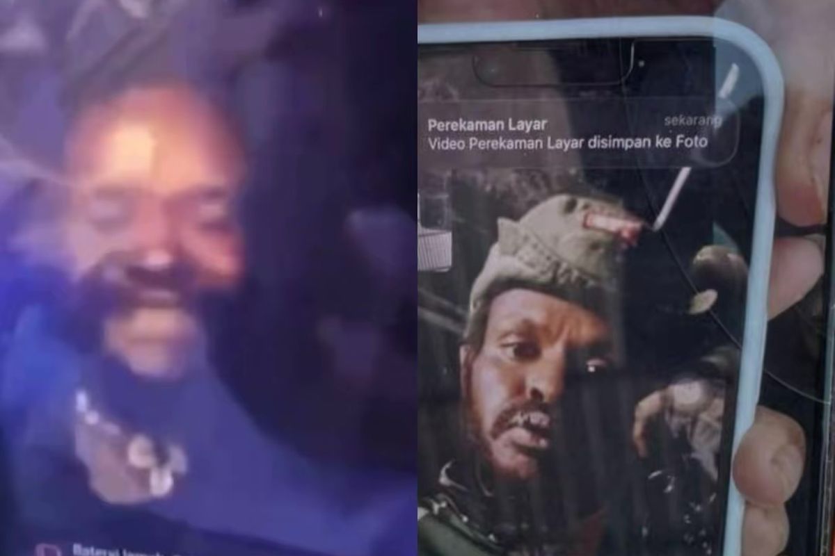 A close-up screenshot of a video call showing an armed militant smiling and laughing while holding a seized mobile phone at a remote outdoor crime scene in Papua.