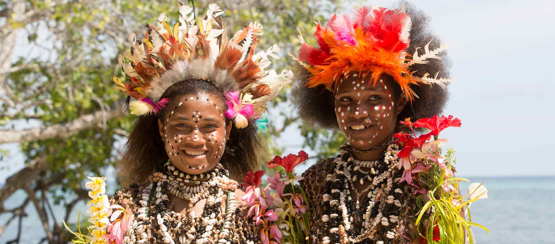 Two young Melanesian girls wearing traditional woven flower crowns smile warmly at the camera outdoors in a lush tropical village.